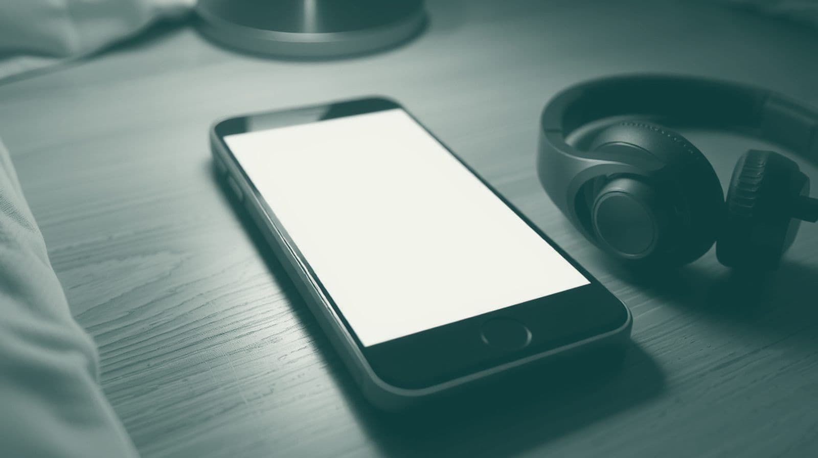 A smartphone with a softly glowing screen on a wooden bedside table at night, beside headphones and a small lamp.