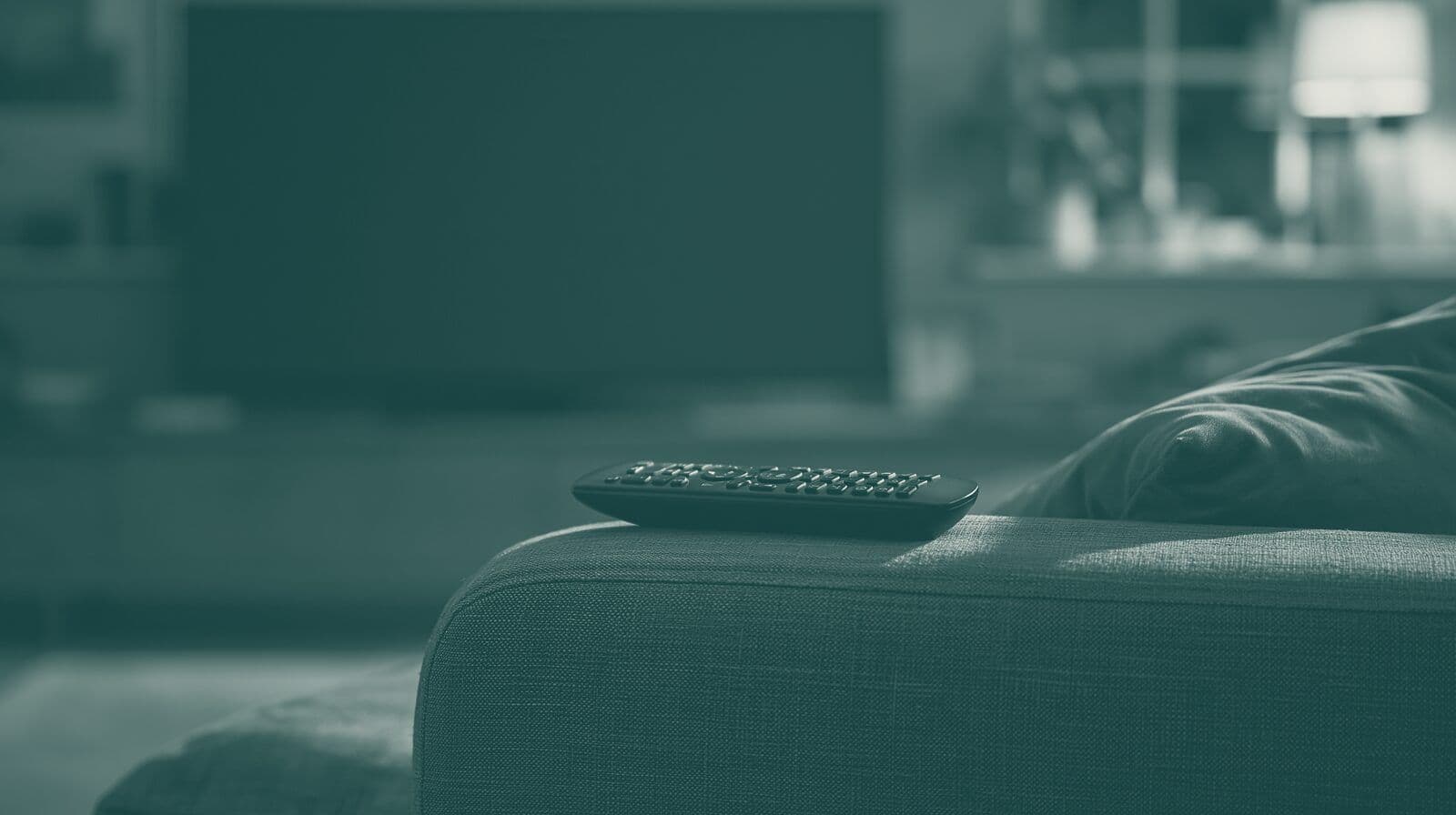 A television remote on the arm of a sofa in front of a dark television.