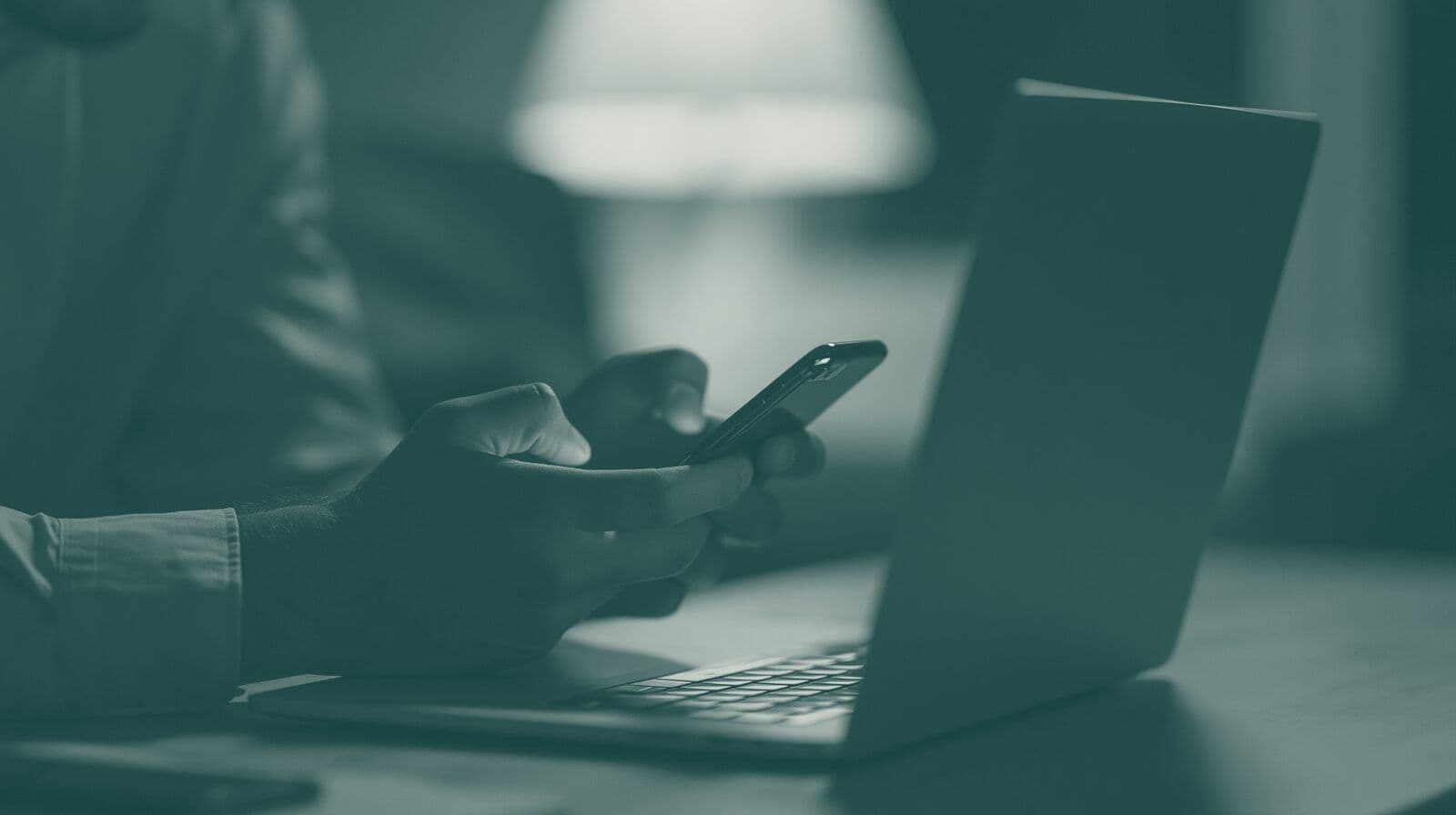Hands hold a phone above an open laptop in a dim room.
