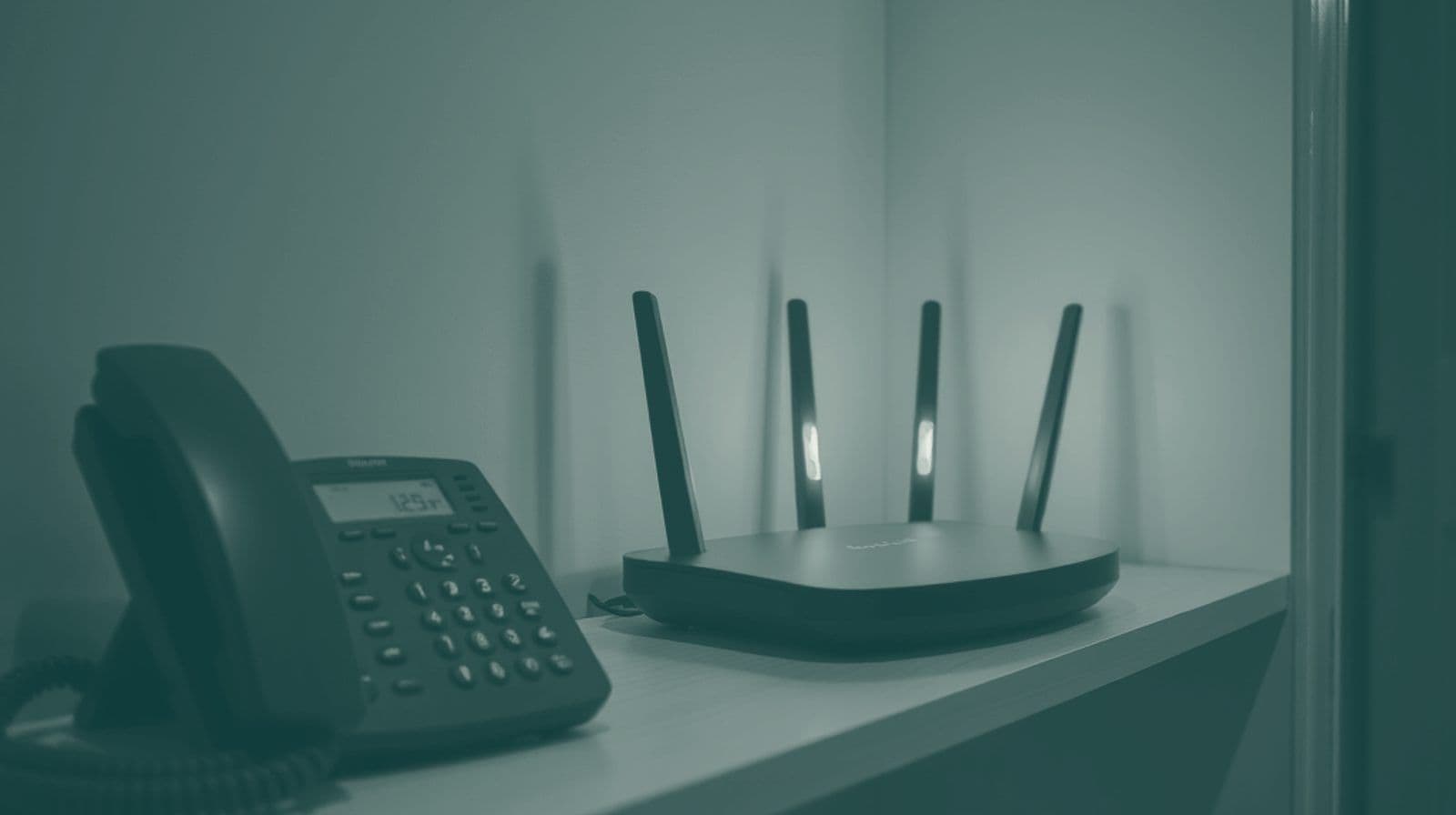 A broadband router with glowing lights on a hallway shelf beside a landline phone, lit from a doorway.